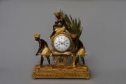 Table Hour: Austrian (?) the end of the 18th century Holder: Gilded and painted figure wooden clock house Dial: enameled, Arabic dial with numbers and varied functions: Structure: Quarter hits.
